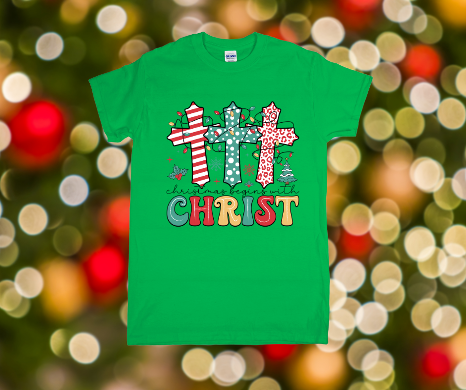 Green t-shirt with Christmas-themed design featuring crosses and text on a blurred festive background.