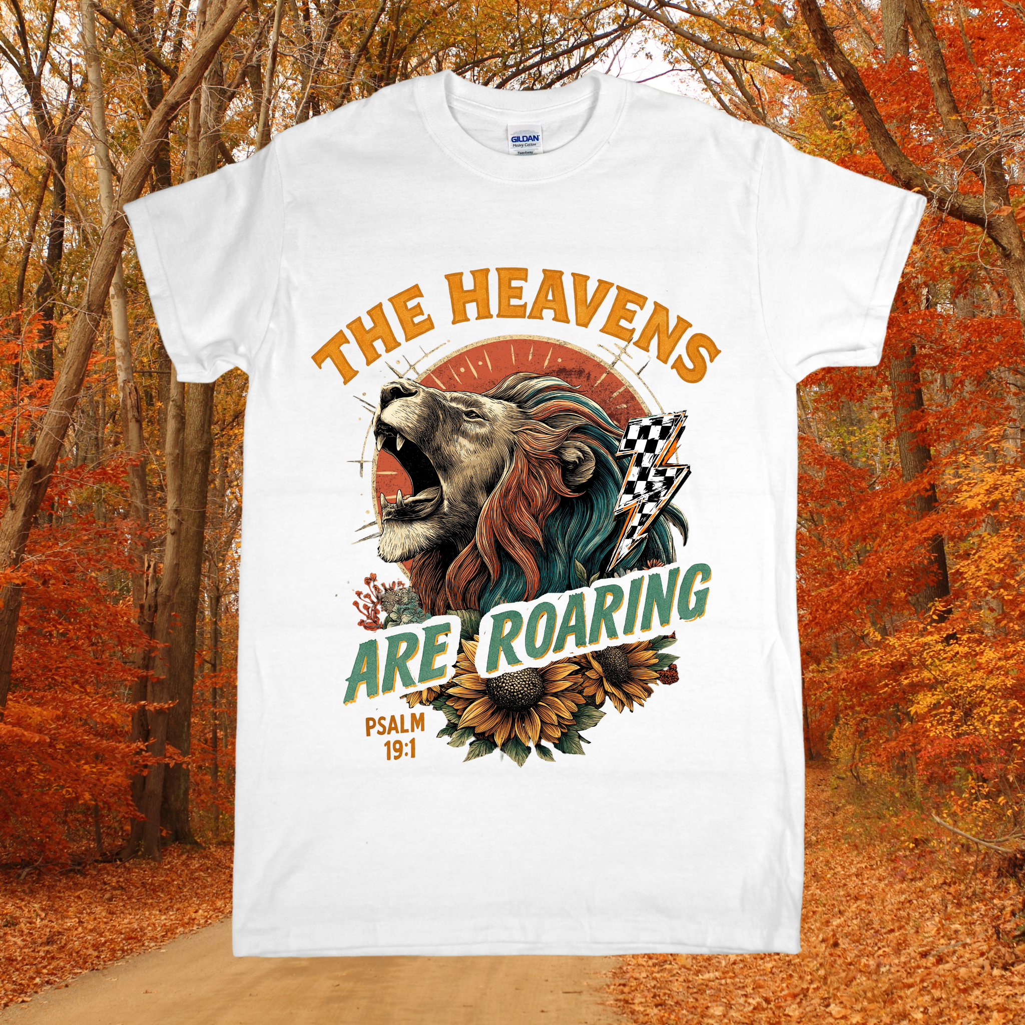 White t-shirt with a graphic design of a lion and sunflower, text 'The Heavens Are Roaring', and Psalm 19:1 reference, set against an autumn forest background.