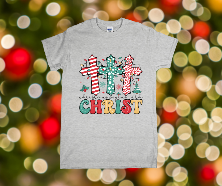 Gray t-shirt with colorful cross design and 'Christmas begins with Christ' text on a festive background.