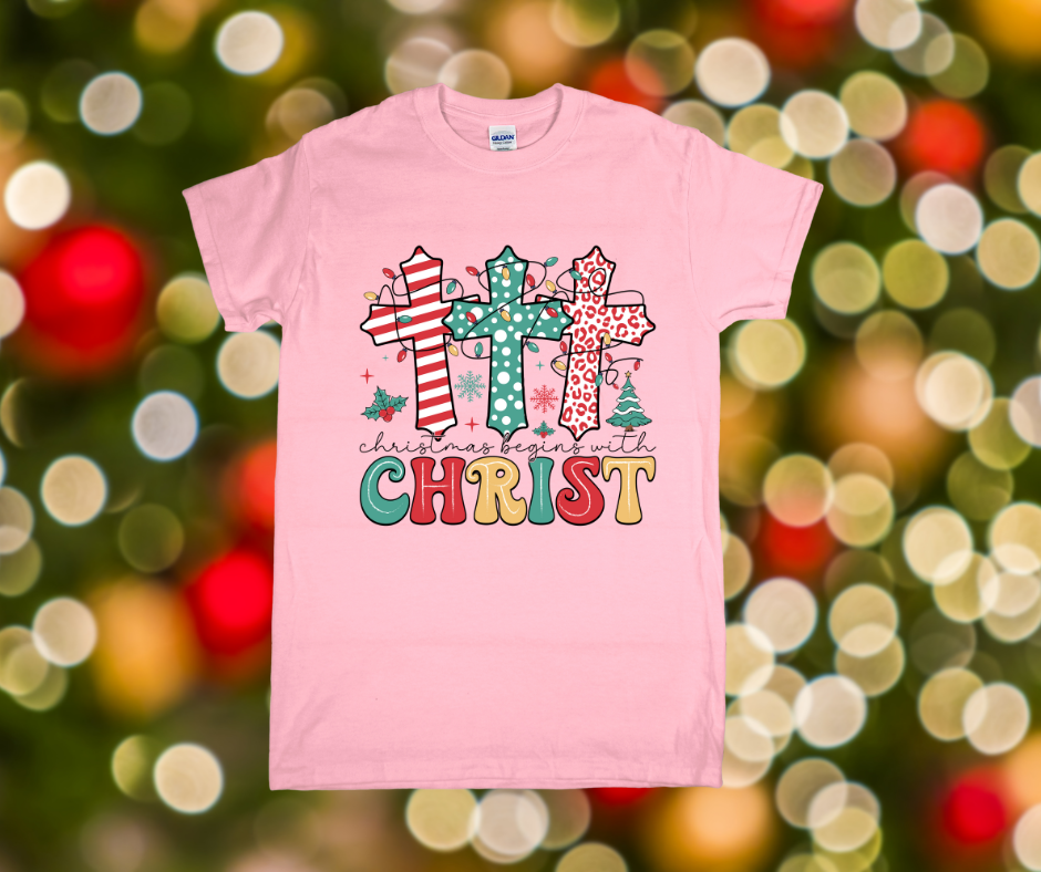 Pink t-shirt with Christmas-themed design featuring crosses and 'Christmas begins with Christ' text on a blurred festive background.
